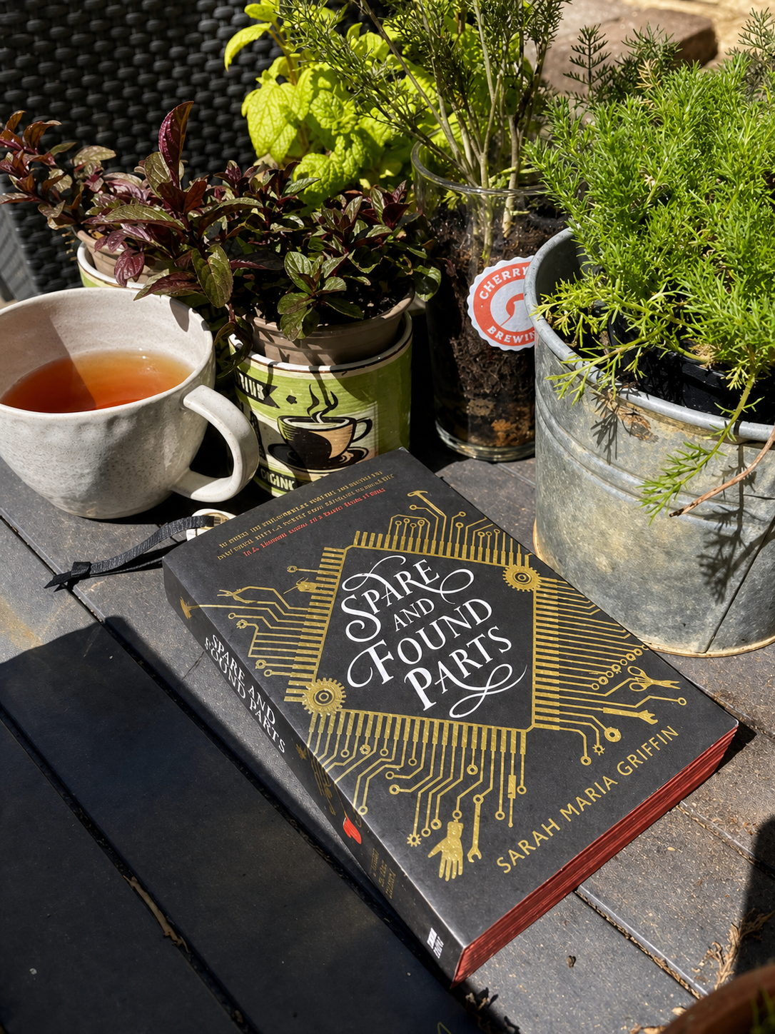 A cup of tea sits beside Spare and Found Parts by Sarah Maria Griffin and potted herbs.